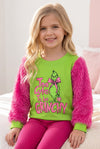 A young girl with blonde hair sits on a bed wearing a festive Grinch-themed pajama set. The set features a bright lime green long-sleeve top with fuzzy hot pink sleeves and matching hot pink flared pants.